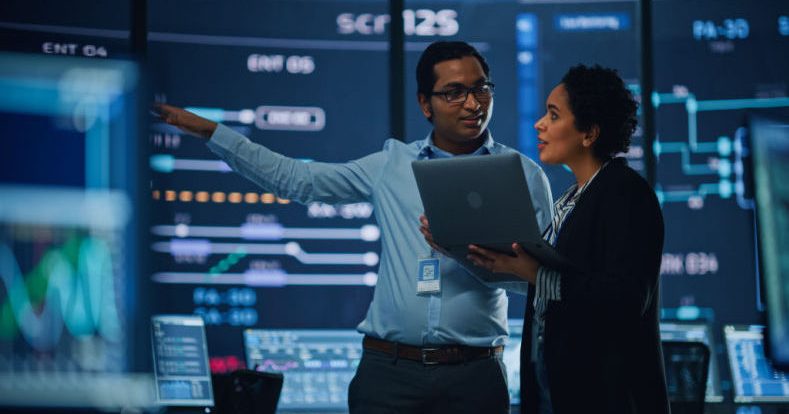 Mann und Frau mit Laptop stehen vor zahlreichen großen Bildschirmen, der Mann scheint etwas zu erklären