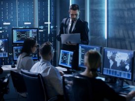 Blick in ein blau beleuchtetes Büro, in dem mehrere Menschen an PCs mit Dashboards zur Netzwerküberwachung sitzen und einem stehenden Mann zuhören, der einen Laptop hält und in diesen blickt