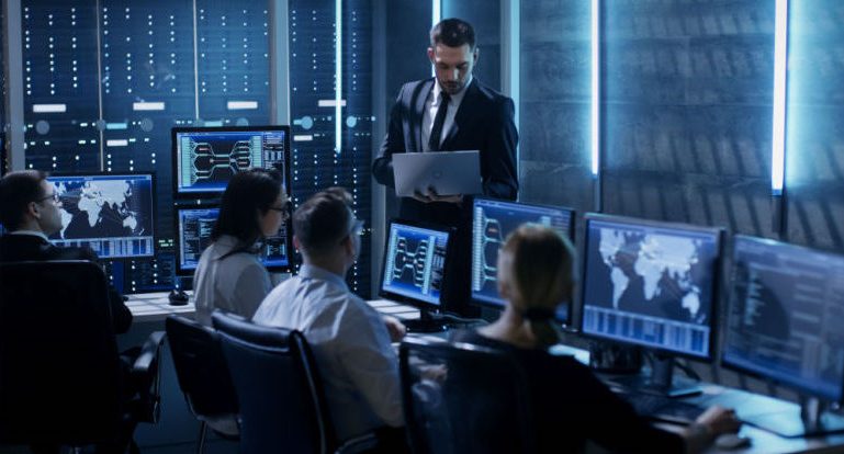 Blick in ein blau beleuchtetes Büro, in dem mehrere Menschen an PCs mit Dashboards zur Netzwerküberwachung sitzen und einem stehenden Mann zuhören, der einen Laptop hält und in diesen blickt