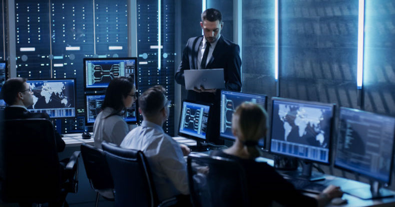 Blick in ein blau beleuchtetes Büro, in dem mehrere Menschen an PCs mit Dashboards zur Netzwerküberwachung sitzen und einem stehenden Mann zuhören, der einen Laptop hält und in diesen blickt