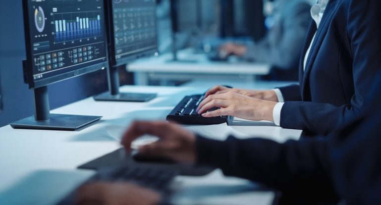 Eine Seitenansicht eines Büroschreibtischs, an dem formal gekleidete Personen vor einem PC-Bildschirm an Tastaturen arbeiten.