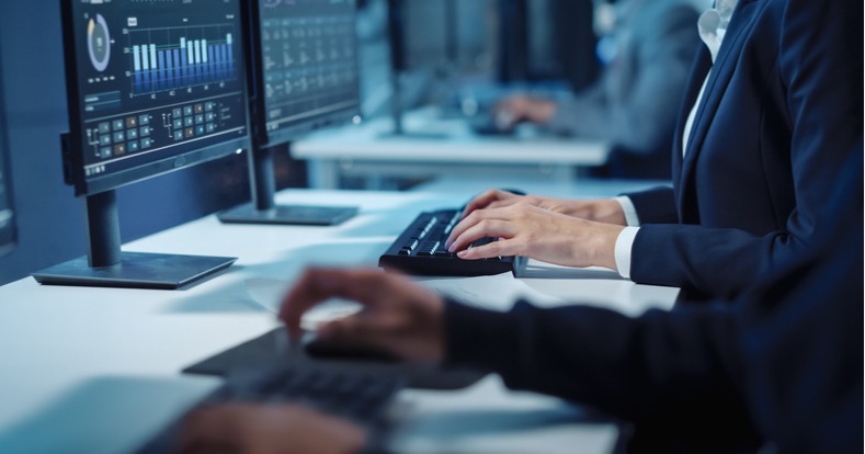 Eine Seitenansicht eines Büroschreibtischs, an dem formal gekleidete Personen vor einem PC-Bildschirm an Tastaturen arbeiten.