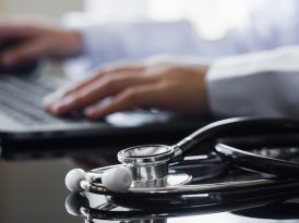 Ein Arzt bedient einen Laptop, vor dem ein Stethoskop liegt.