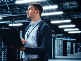 Ein Mann steht mit einem Laptop in der Hand in einem Serverraum.